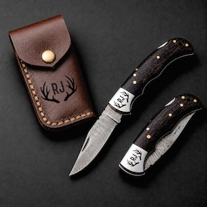 May include: Two folding knives with dark wood handles, brass pins, and Damascus steel blades, alongside a brown leather sheath. The knives and sheath feature a laser-engraved logo with the letters RJ flanked by two deer antler silhouettes.