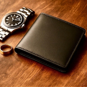 Black leather bifold wallet on a warm wooden surface with a silver wristwatch and gold ring nearby, creating a classy, masculine, gift-ready presentation.