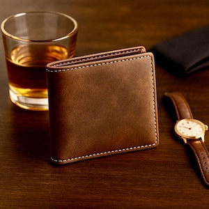 Brown leather bifold wallet displayed on a dark wooden surface with a glass of whiskey and a brown-band wristwatch in the background, creating a warm, classy, masculine aesthetic.