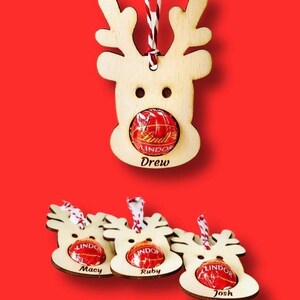 May include: Four wooden reindeer ornaments with a red candy inside each. The reindeer have names written below them: Drew, Macy, Ruby, and Josh.