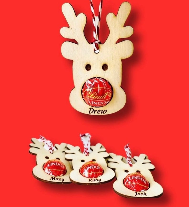 May include: Four wooden reindeer ornaments with a red candy inside each. The reindeer have names written below them: Drew, Macy, Ruby, and Josh.