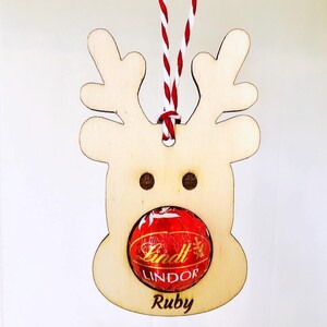May include: Wooden reindeer ornament with a red and white twine hanger. The reindeer has a red Lindt Lindor chocolate in its mouth and the name "Ruby" is written below the chocolate.