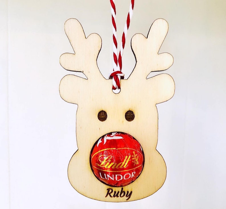 May include: Wooden reindeer ornament with a red and white twine hanger. The reindeer has a red Lindt Lindor chocolate in its mouth and the name "Ruby" is written below the chocolate.