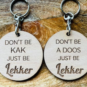 Puede incluir: Dos llaveros de madera con anillas de plata. Los llaveros son redondos y tienen el texto "Don't be kak just be lekker" y "Don't be a doos just be lekker" grabado en ellos.