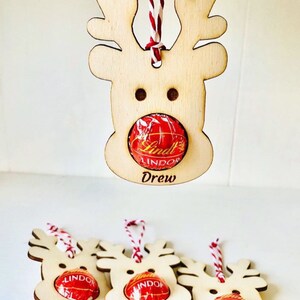 May include: Four wooden reindeer ornaments with red and white twine. Each ornament has a space for a candy, and the names Drew, Macy, Ruby, and Josh are written below the candy spaces.