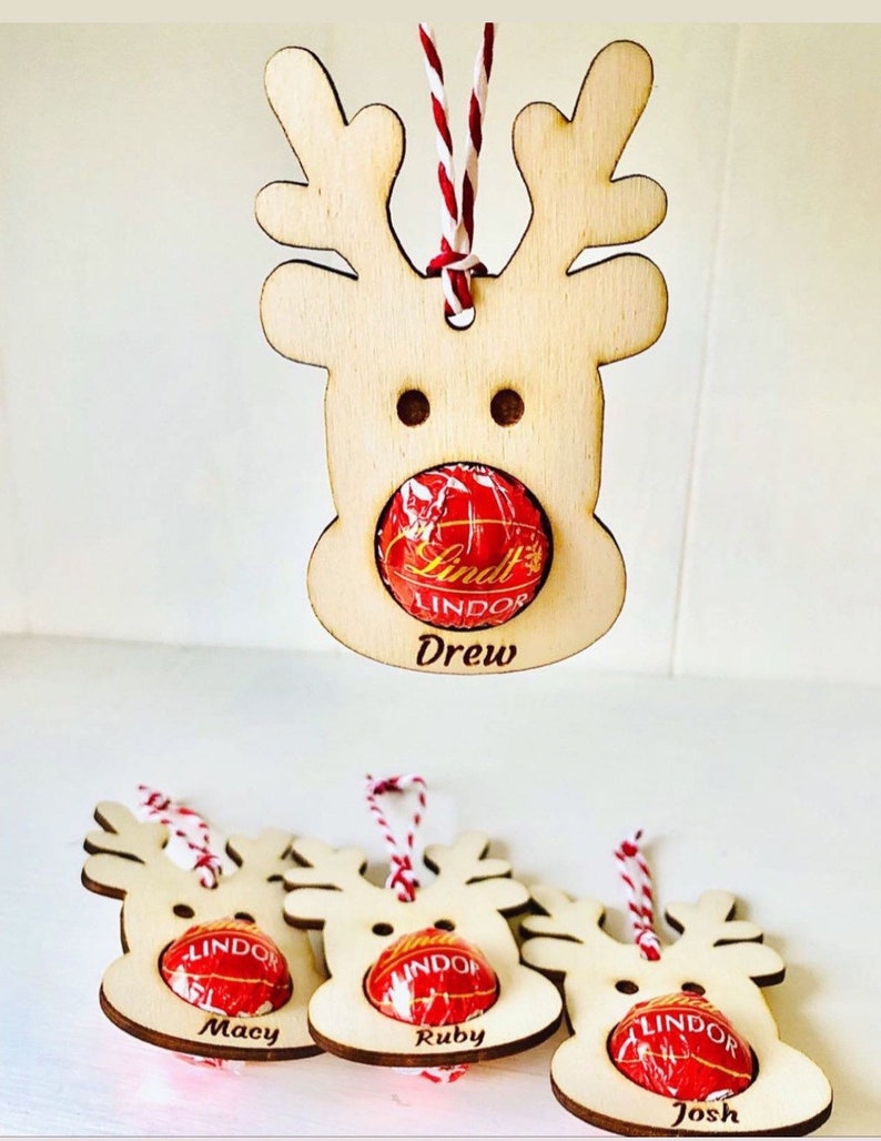 May include: Four wooden reindeer ornaments with red and white twine. Each ornament has a space for a candy, and the names Drew, Macy, Ruby, and Josh are written below the candy spaces.