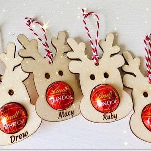 May include: Four wooden reindeer ornaments with red and white twine hangers. Each ornament has a Lindor chocolate in the center and a name written below: Drew, Macy, Ruby, and Josh.
