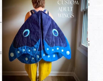 May include: A person wearing custom adult wings. The wings are a deep blue with lighter blue accents and white spots. The text "CUSTOM ADULT WINGS" is visible in the image.