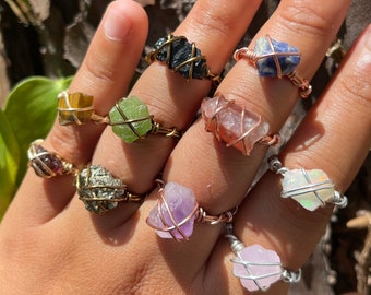 Puede incluir: Una colección de nueve anillos envueltos en alambre con piedras preciosas en bruto y sin tallar en varios colores, incluyendo negro, azul, verde, amarillo, oro metálico, morado, rosa y blanco iridiscente, asegurados con bandas de metal.
