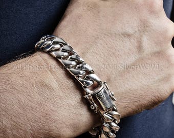 May include: A silver curb chain bracelet with a secure clasp. The bracelet is made of interlocking links and is displayed on a person's wrist. The clasp has a detailed design. The bracelet is a piece of jewelry.
