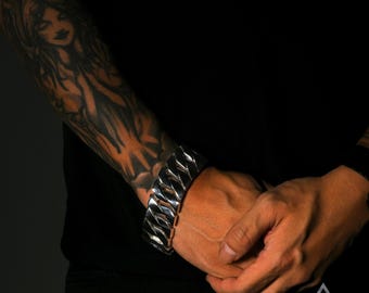 May include: A silver chain bracelet with a large, chunky link design. The bracelet is worn on the wrist of a person with a tattoo on their arm.