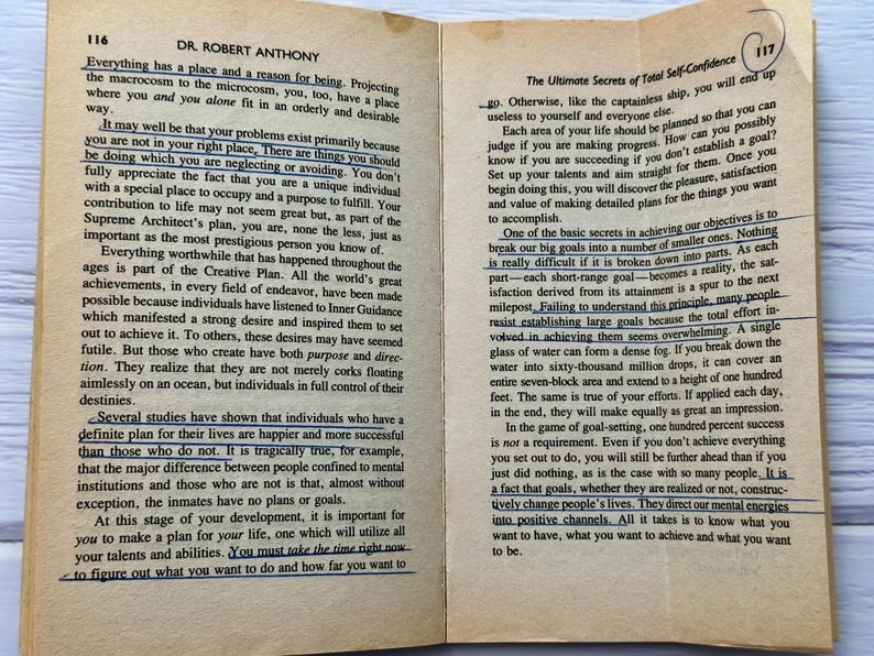 May include: An open book with text on aged paper. The pages are from a book titled "Dr. Robert Anthony" and "The Ultimate Secrets of Total Self-Confidence". The text is highlighted with blue ink. The book is open to pages 116 and 117.