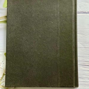 May include: A hardcover book with a dark olive green cover. The spine is a slightly darker shade of green. The book is rectangular and appears to be closed. White flowers are in the background.