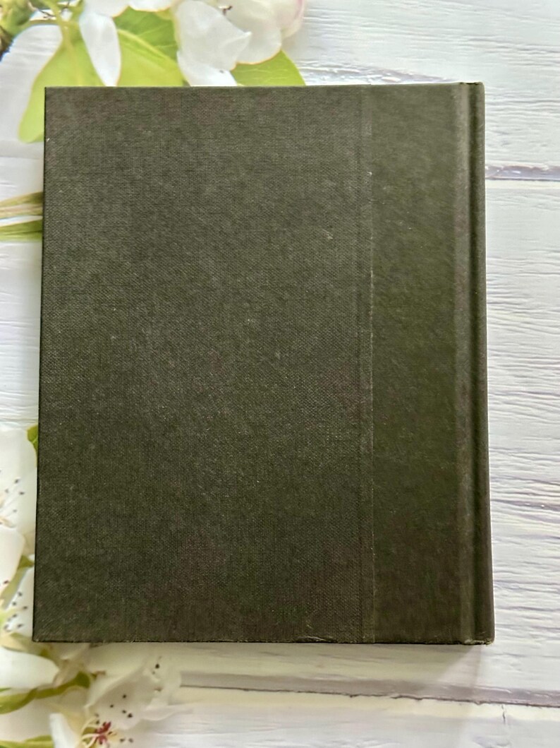 May include: A hardcover book with a dark olive green cover. The spine is a slightly darker shade of green. The book is rectangular and appears to be closed. White flowers are in the background.
