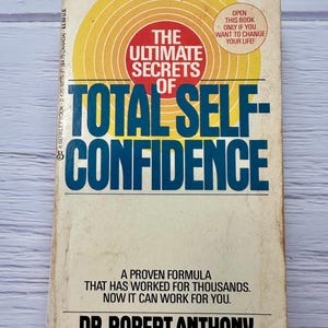 May include: A well-worn paperback book with the title "The Ultimate Secrets of Total Self-Confidence" by Dr. Robert Anthony. The cover design includes a red circle, yellow concentric circles, and the text "Open this book only if you want to change your life!"