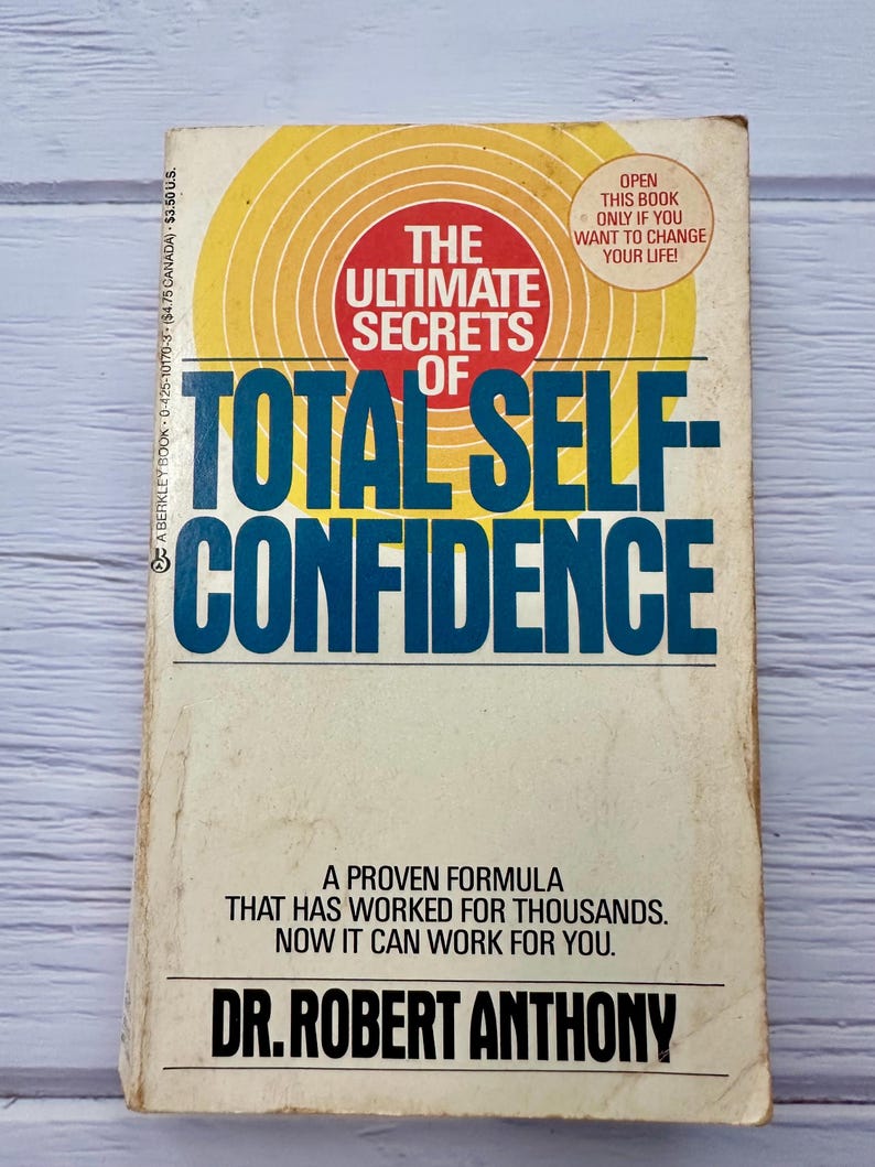 May include: A well-worn paperback book with the title "The Ultimate Secrets of Total Self-Confidence" by Dr. Robert Anthony. The cover design includes a red circle, yellow concentric circles, and the text "Open this book only if you want to change your life!"