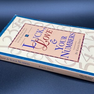 May include: A beige book titled "Luck, Love & Your Numbers" by Karen David. The cover features a background of large numbers and the title is written in a decorative font.