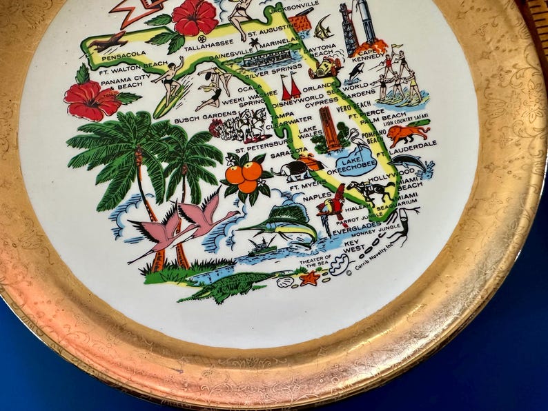 Pode incluir: Um prato decorativo com borda dourada e centro branco com um mapa colorido da Fl&oacute;rida. O mapa inclui ilustra&ccedil;&otilde;es de palmeiras, flamingos e v&aacute;rios marcos e cidades da Fl&oacute;rida.