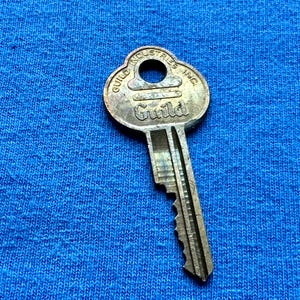 May include: A vintage metal key with the word "Guild" and "Guild Industries Inc." engraved on it. The key is silver-colored and has a unique shape with a circular hole at the top. The key is set against a blue fabric background.