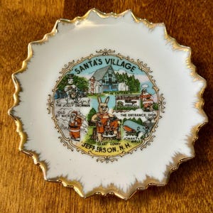 May include: A small white porcelain decorative plate with a scalloped gold-trimmed edge. The center features a colorful illustration of Santa's Village in Jefferson, N.H., depicting a snowman, a giant bunny, Old St. Nick, and a whale.