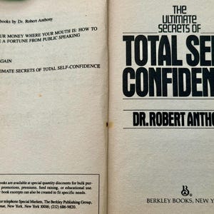 May include: Open book with the title "The Ultimate Secrets of Total Self-Confidence" by Dr. Robert Anthony. The left page lists related titles including "Put Your Money Where Your Mouth Is". Published by Berkley Books, New York.