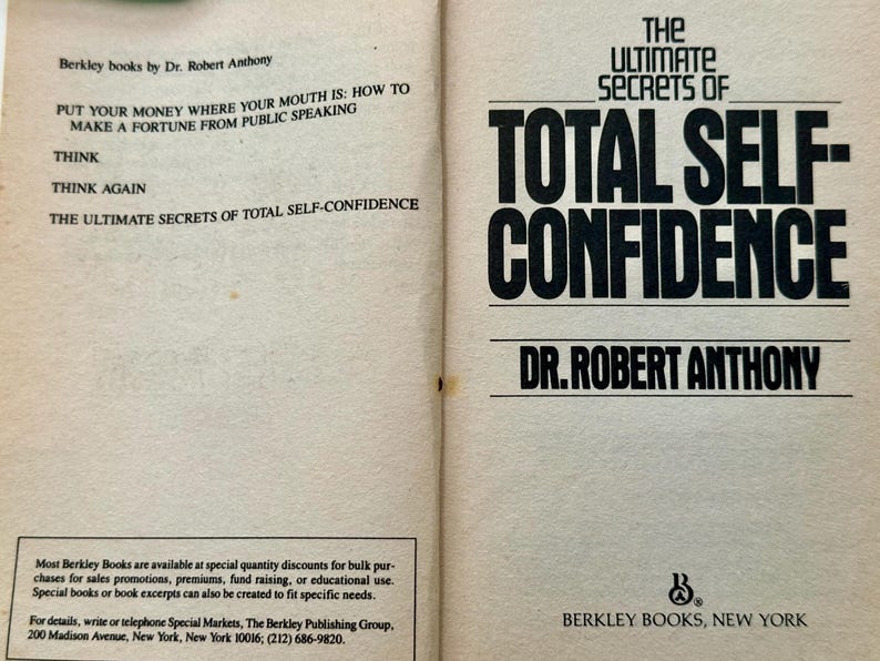 May include: Open book with the title "The Ultimate Secrets of Total Self-Confidence" by Dr. Robert Anthony. The left page lists related titles including "Put Your Money Where Your Mouth Is". Published by Berkley Books, New York.