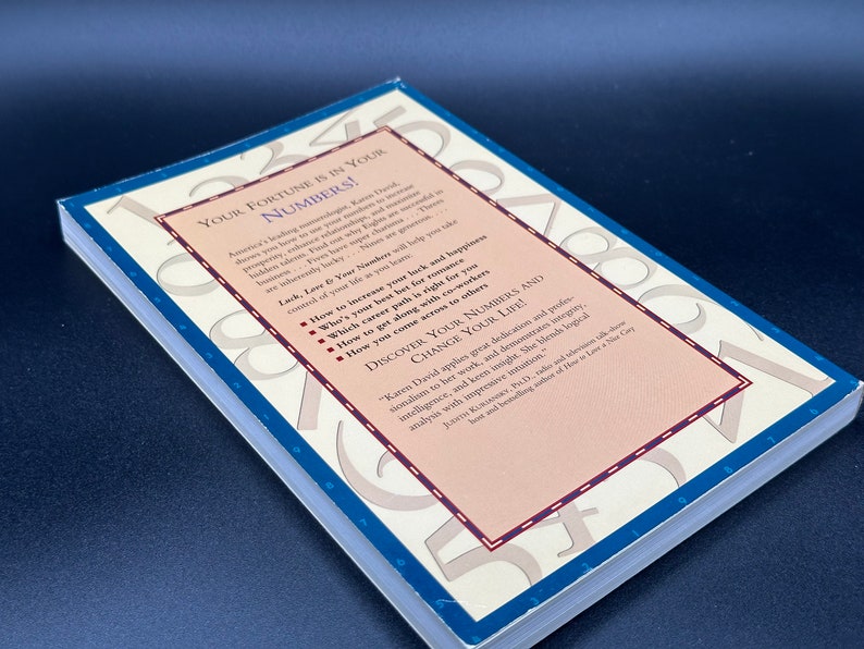 May include: A book cover with a pink and white background. The title of the book is "Your Fortune is in Your Numbers!" The book cover features a variety of numbers in a circular pattern. The book cover also includes a description of the book, which includes information about how to increase your luck and happiness, how to find your best career path, and how to get along with colleagues.