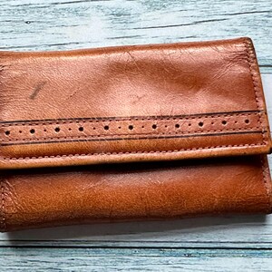 May include: A brown leather wallet with a decorative stitch detail. The wallet has a fold-over closure and a smooth, worn texture.