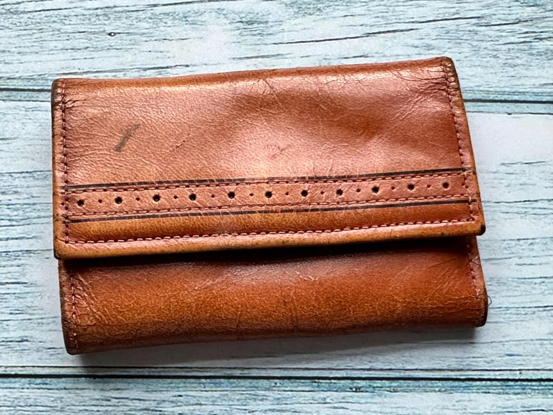 May include: A brown leather wallet with a decorative stitch detail. The wallet has a fold-over closure and a smooth, worn texture.