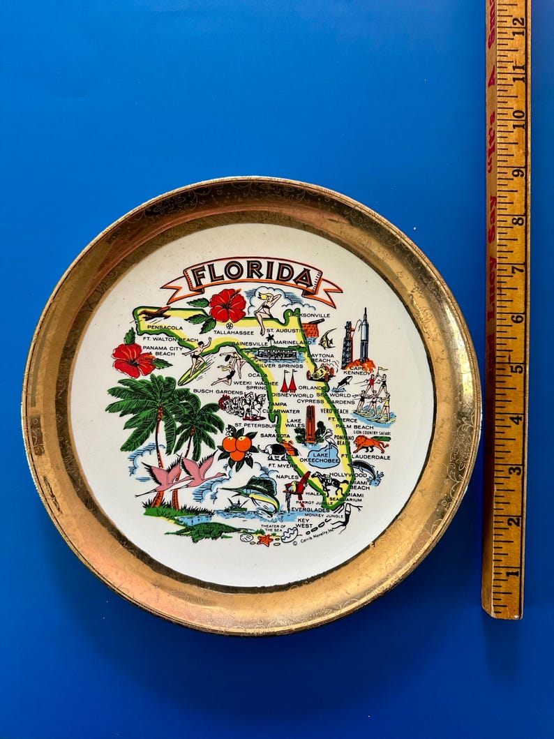 Pode incluir: Um prato decorativo com uma borda dourada com um mapa colorido da Fl&oacute;rida. O mapa inclui nomes de cidades, pontos de refer&ecirc;ncia e ilustra&ccedil;&otilde;es de palmeiras e flores. O prato tem aproximadamente 15 cm de di&acirc;metro.