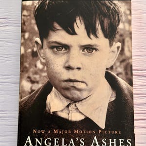 May include: A hardcover book titled "Angela's Ashes" by Frank McCourt. The cover features a sepia-toned portrait of a young boy. Text includes "The #1 New York Times Bestseller" and "Now a Major Motion Picture."