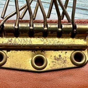 May include: Close-up of a gold-toned metal clasp with the words "HANNA" and "U.S.I.P.A. 26482" engraved on it. The clasp is attached to a brown leather surface.