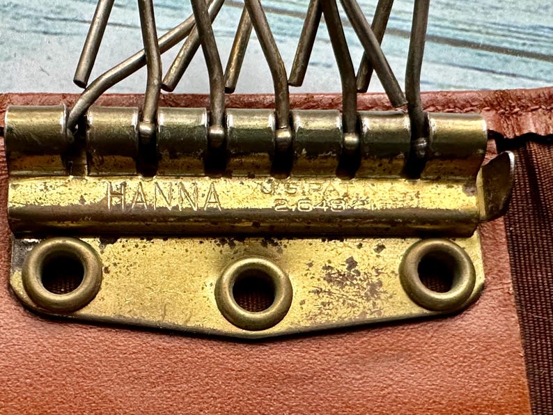 May include: Close-up of a gold-toned metal clasp with the words "HANNA" and "U.S.I.P.A. 26482" engraved on it. The clasp is attached to a brown leather surface.