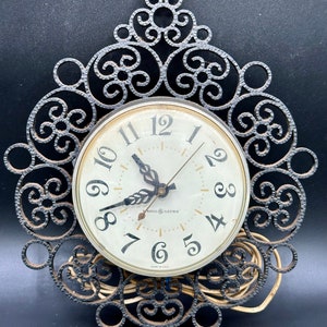 May include: A vintage General Electric electric clock with a white face and black hands. The clock is mounted in a black metal frame with a decorative scroll design. The clock is made in the USA.