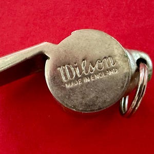 May include: A silver whistle with a black inscription that reads "Wilson Made in England".