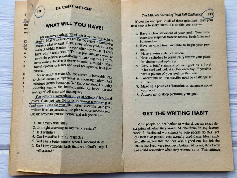 May include: Open book pages with text on self-confidence and goal setting. The text includes questions to ask about goals and steps to achieve them. The pages are aged with a yellowed tone.