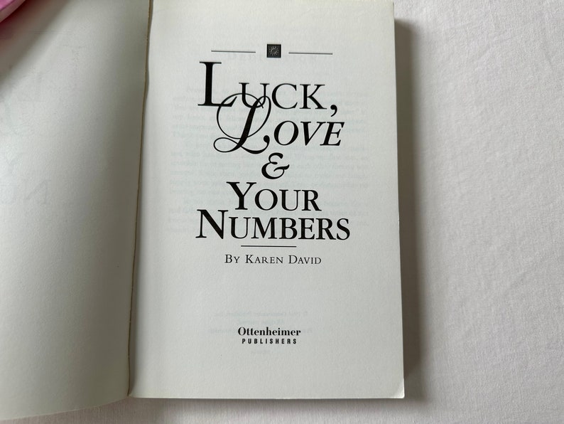 May include: A white book with the title "Luck, Love & Your Numbers" by Karen David. The book is published by Ottenheimer Publishers.