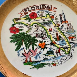 Pode incluir: Um prato decorativo vintage com uma borda dourada e um centro branco com um mapa colorido da Fl&oacute;rida. O mapa inclui ilustra&ccedil;&otilde;es de marcos, cidades e s&iacute;mbolos estaduais, como flores de hibisco e palmeiras.