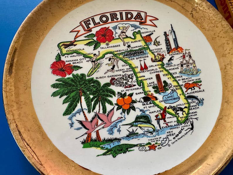 Pode incluir: Um prato decorativo vintage com uma borda dourada e um centro branco com um mapa colorido da Fl&oacute;rida. O mapa inclui ilustra&ccedil;&otilde;es de marcos, cidades e s&iacute;mbolos estaduais, como flores de hibisco e palmeiras.