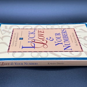May include: A book titled "Luck, Love & Your Numbers" by Karen David. The cover features a pink and white background with a large number "1" in the background. The book is about numerology and how to change your life.