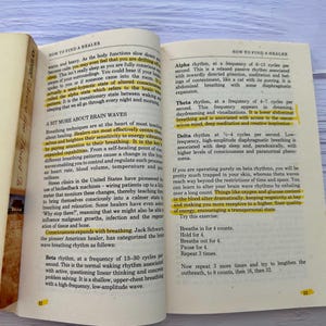 May include: Open book with text about brain waves and healing techniques. The pages are yellowed, with highlighted sections in yellow. The book is open on a light blue and white surface.