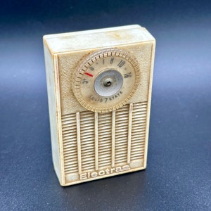Puede incluir: Una radio de transistores Electra 7-State vintage de color blanco con un dial redondo y una rejilla de altavoz. La radio tiene una carcasa de plástico de color crema y una perilla negra.