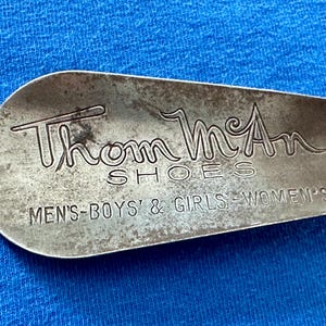 May include: A vintage metal shoehorn with the text "Thom McAn SHOES" and the words "MEN'S-BOYS' & GIRLS-WOMEN'S" in a serif font. The shoehorn has a worn, aged appearance.