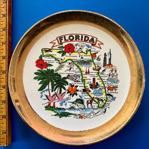 Pode incluir: Um prato decorativo vintage com um mapa da Fl&oacute;rida com ilustra&ccedil;&otilde;es de marcos e flora. O prato tem uma borda dourada e a palavra "FLORIDA" em uma faixa. O prato mede aproximadamente 15 cm de di&acirc;metro.