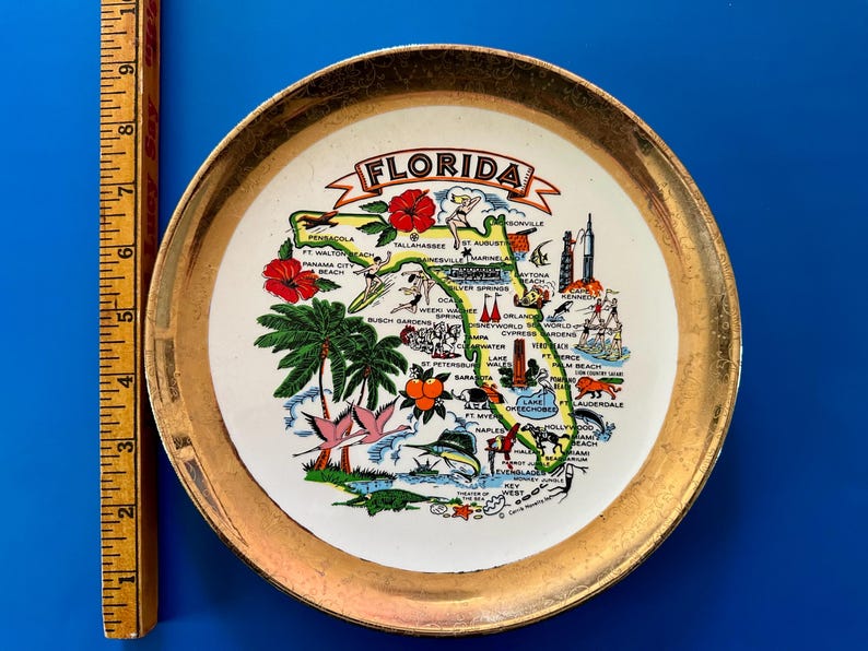 Pode incluir: Um prato decorativo vintage com um mapa da Fl&oacute;rida com ilustra&ccedil;&otilde;es de marcos e flora. O prato tem uma borda dourada e a palavra "FLORIDA" em uma faixa. O prato mede aproximadamente 15 cm de di&acirc;metro.