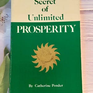 May include: A vintage book titled "Secret of Unlimited Prosperity" by Catherine Ponder. The book has a cream-colored top and a green bottom. The cover features a gold sunburst design.