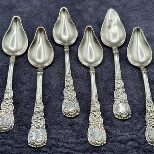 May include: A set of six silver-toned metal spoons featuring teardrop-shaped bowls and ornate, embossed floral and scrollwork detailing along the handles. Each spoon has a consistent decorative pattern on the stem and a smooth, polished finish.