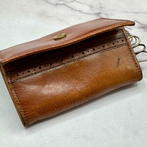 May include: A brown leather key holder with a decorative stitch pattern and a metal key ring. The key holder is closed and has a flap.