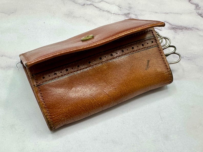 May include: A brown leather key holder with a decorative stitch pattern and a metal key ring. The key holder is closed and has a flap.