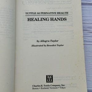 May include: Open book with the title "Healing Hands" by Allegra Taylor. The book is published by Charles E. Tuttle Company, Inc. The cover is off-white with black text. The book is open to the title page.